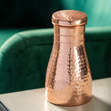 A hammered copper shaker bottle with a matching lid, featuring a floral motif embossed on the lower front, set on a light surface with a green velvet chair in the background.