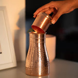 A hand pouring liquid from a small hammered copper cup into a larger matching copper vessel on a tabletop in warm indoor lighting.