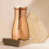 A hammered copper tumbler with a matching lid, styled on a dark stone block with a light stone wedge behind it against a warm beige background.