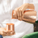 A person in a white shirt pouring clear liquid from a hammered copper pitcher into a small copper tumbler, with a green chair visible in the background.