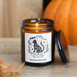 Candle in an amber jar with a decorative label featuring a cat design, on a wooden surface with a blurred pumpkin in the background.