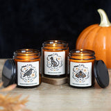 Three lit candles in amber jars with decorative labels on a wooden surface, accompanied by a pumpkin.