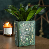 Green Witch book of spells with a potted plant and lit candle on a wooden surface