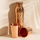 A copper-toned hammered metal water bottle and two matching cups arranged on a warm off-white surface with soft shadows.