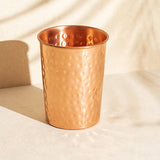A hammered copper tumbler with a polished metallic finish, standing on a light beige surface with soft shadows cast across it.