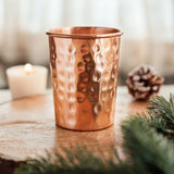 A hammered copper tumbler cup sits on a rustic wooden slice with a lit candle, pinecone, and evergreen branches in a softly blurred indoor setting.