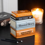 Stack of book-shaped containers with titles on a dark surface, lit by a candle.
