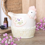 Ceramic chicken-shaped container with floral design on a wooden surface with flowers and a bottle in the background.