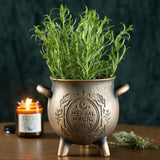 Bronze pot labeled 'Herbal Magic' with a plant, candle, and lavender on a wooden surface.