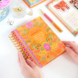 Colorful notebook with floral design held by a hand on a white surface with stationery items.