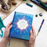 Blue decorative journal held by hands on a white surface with a pen and small objects.