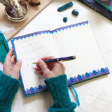 Person writing in a decorated notebook with a pen on a white surface.