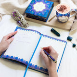 Person writing in a notebook with a decorative cover, surrounded by a mug, box, and small plants on a white surface.