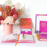 Colorful stationery items including notebooks, a pen, and a floral arrangement on a white surface.