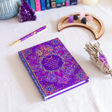 Purple notebook with decorative cover on a white surface with crystals and candles