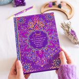 Purple journal with mandala design held by a person on a white surface with decorative items.