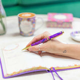 Hand holding a decorative pen over an open notebook with a blurred coffee cup and stationery in the background.
