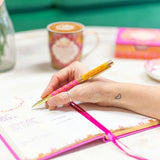 Person holding a decorative pen over an open notebook with a cup of coffee in the background.