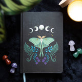 Black notebook with a green moth illustration on a dark surface with plants and candles.