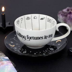 White mug with 'telling fortunes in tea' text on a black saucer with star designs, against a dark background.