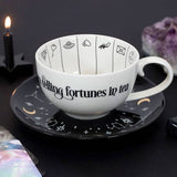 White mug with 'telling fortunes in tea' text on a black saucer with star designs, against a dark background.