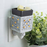 Wall-mounted wax warmer with decorative design on a tiled wall next to a plant.