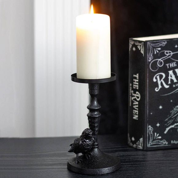 Black candle holder with a lit white candle next to a book titled 'The Raven' on a dark surface.