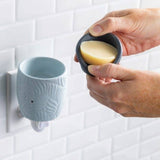 Person holding a bar of soap in a blue soap dish against a tiled wall.