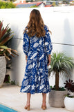 Woman in a blue floral dress standing on a balcony with plants and a pool view.