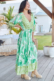 Woman in a green and white patterned dress standing on a wooden deck with plants and furniture in the background.