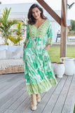 Woman in a green and white patterned dress standing on a wooden deck with outdoor furniture in the background.