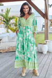 Woman wearing a green patterned dress standing on a wooden deck with plants and furniture in the background.