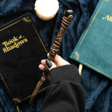Hand holding a decorative wand with 'Book of Shadows' and green book in the background