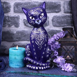 Decorative purple cat figurine with mystical symbols next to a lit candle and flowers against a stone wall.