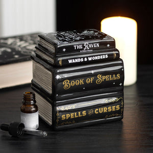 Stack of books titled 'The Raven', 'Wands & Wonders', and 'Book of Spells' with a small bottle on a dark surface.