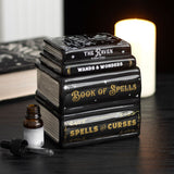 Stack of books titled 'The Raven', 'Wands & Wonders', and 'Book of Spells' with a small bottle on a dark surface.