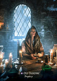 Person in a hooded robe sitting at a table with candles and books in a dimly lit room.