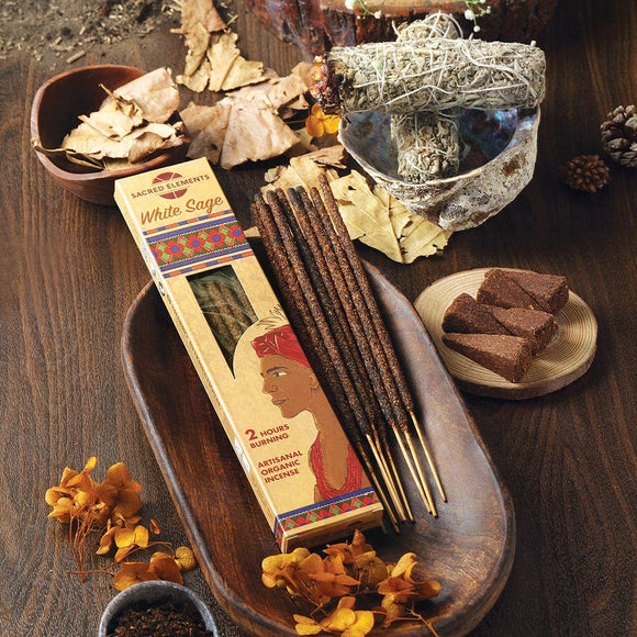 White sage incense sticks with packaging on a wooden tray, surrounded by dried herbs and elements.