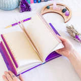 Open notebook with purple binding held by a hand on a white surface with decorative items.