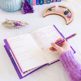 Person writing in a purple planner with crystals and decor on a white surface