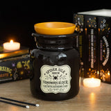 A black glass jar candle labeled 'Spider Silk Serum' and 'Venomous Klyster' with a yellow wax dish on top, styled on a tabletop with candles and books in the background.