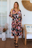 Woman wearing a floral dress standing in a room with a chair and decorative items.