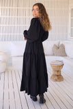 Woman in a black dress standing on a wooden deck with a white wall and cushioned sofa in the background.
