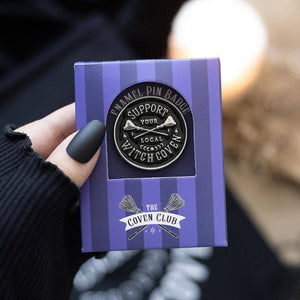 Enamel pin badge in a purple striped box held by a hand with dark nail polish.
