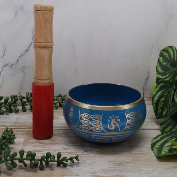 Blue singing bowl with gold patterns, wooden handle, and red cushion on a marble surface with green plants.