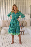 Woman wearing a green patterned dress standing in a room with white walls and wooden floor.