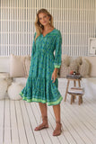 Woman wearing a green dress in an outdoor setting with a white wall and wooden floor.