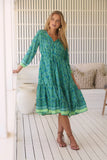 Woman wearing a green patterned dress standing in a bright room with white walls and wooden floor.