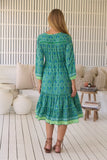 Woman wearing a green patterned dress standing in a room with white walls and wooden floor.