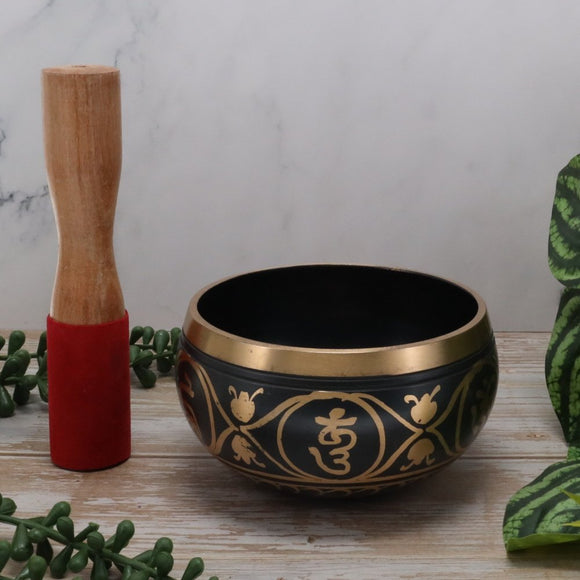 Black singing bowl with gold designs and a wooden mallet on a light surface.